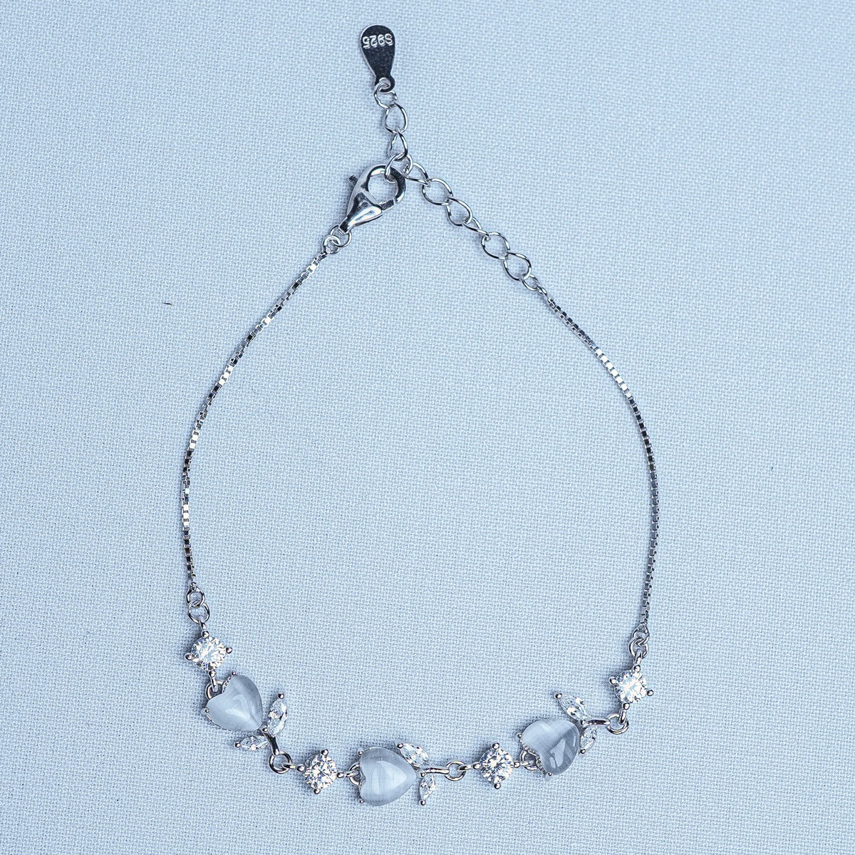 A silver chain bracelet featuring a row of light blue-gray teardrop-shaped crystals and small clear cubic zirconia accents, displayed on a light blue textured background.